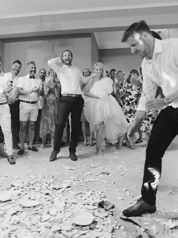 Groom dances joyfully in white shirt, holding his head laughing — bride in lace beside him, confetti on floor