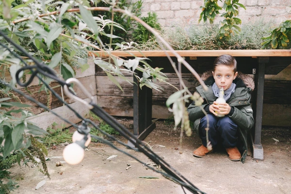 Boy crouches at a wooden structure among green plants holding an apple — rural wedding setting