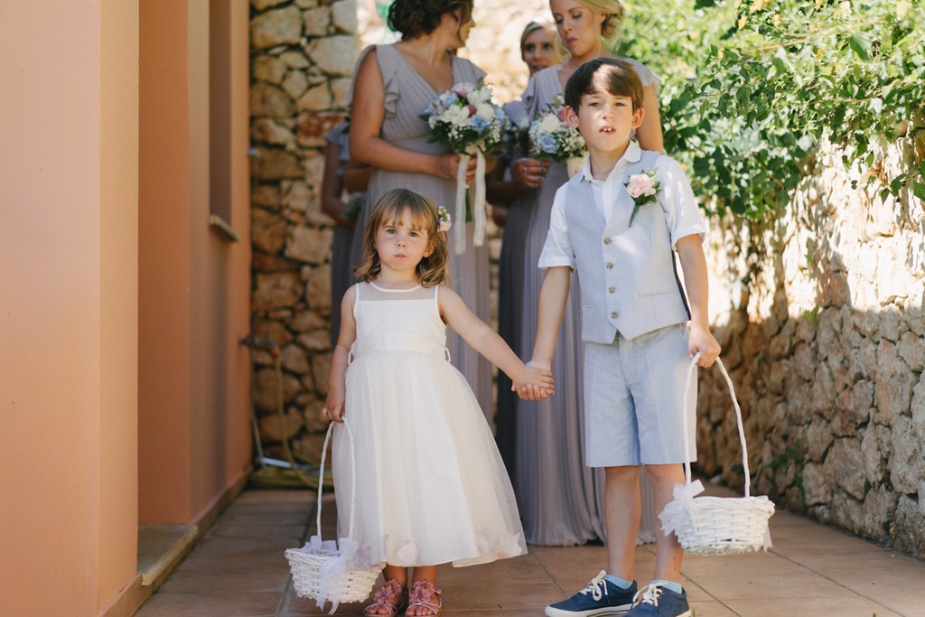 Two children walk hand in hand scattering white flowers — girl in white dress, boy in light suit