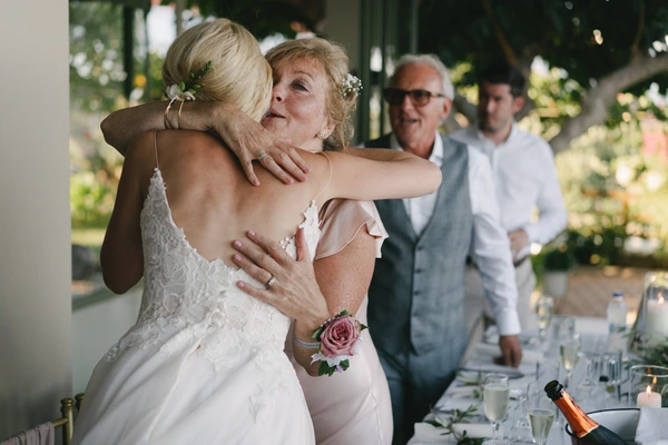 Bride in white lace welcomed with a warm embrace by an older woman, guests smiling in a green garden