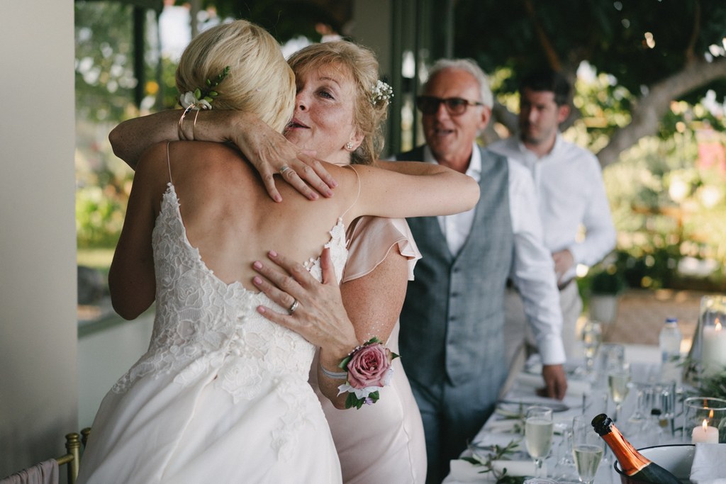 Bride in white lace welcomed with a warm embrace by an older woman, guests smiling in a green garden