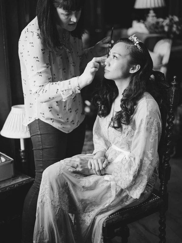 Makeup artist works on the bride, who sits patiently in white lace with a silver headpiece