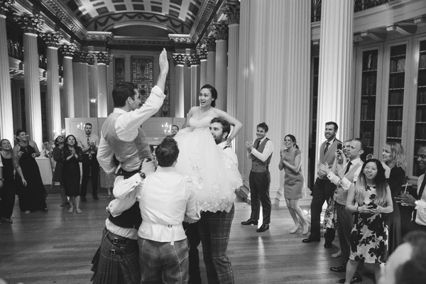 Bride lifted in the air by guests mid-dance in a classical hall — black-and-white, joyful moment