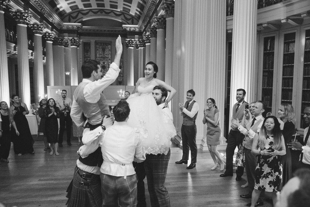 Bride lifted in the air by guests mid-dance in a classical hall — black-and-white, joyful moment