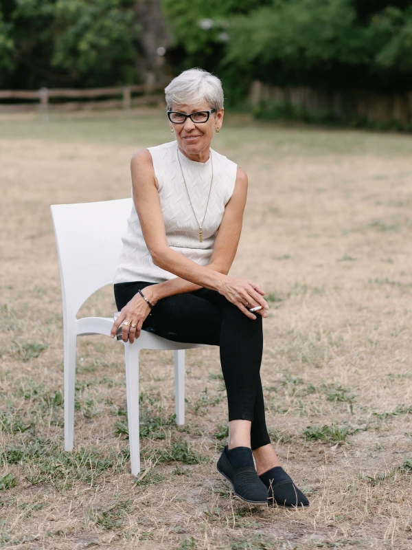 Elderly woman with glasses smiling sitting on white chair at outdoor wedding venue in natural setting.