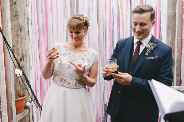 Bride and groom smiling with whisky glasses — cream lace top, tulle skirt, navy suit