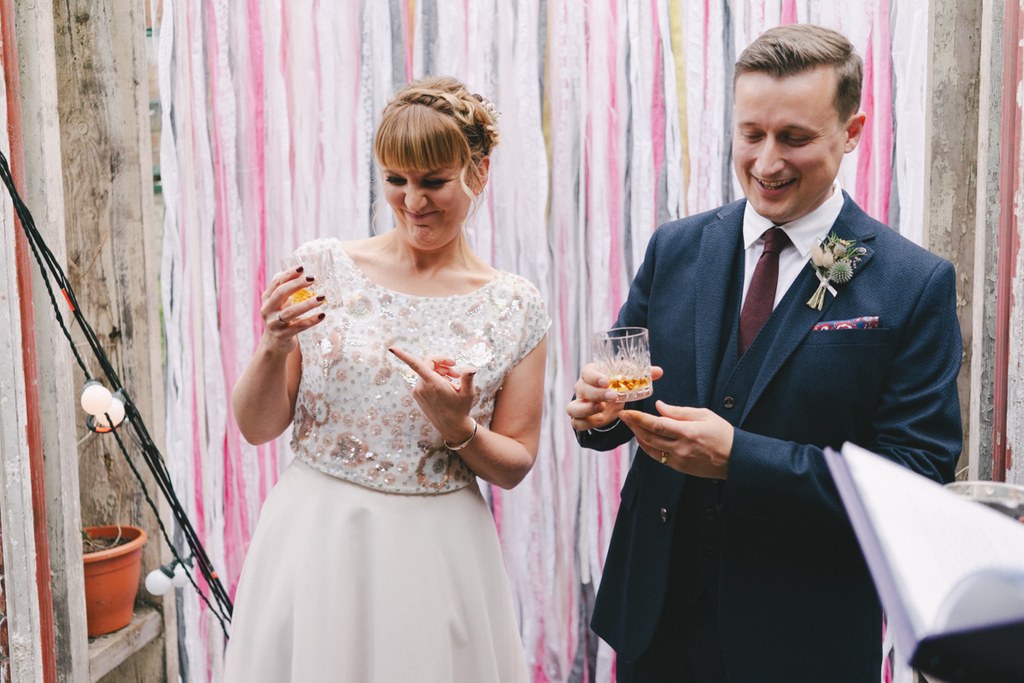 Bride and groom smiling with whisky glasses — cream lace top, tulle skirt, navy suit