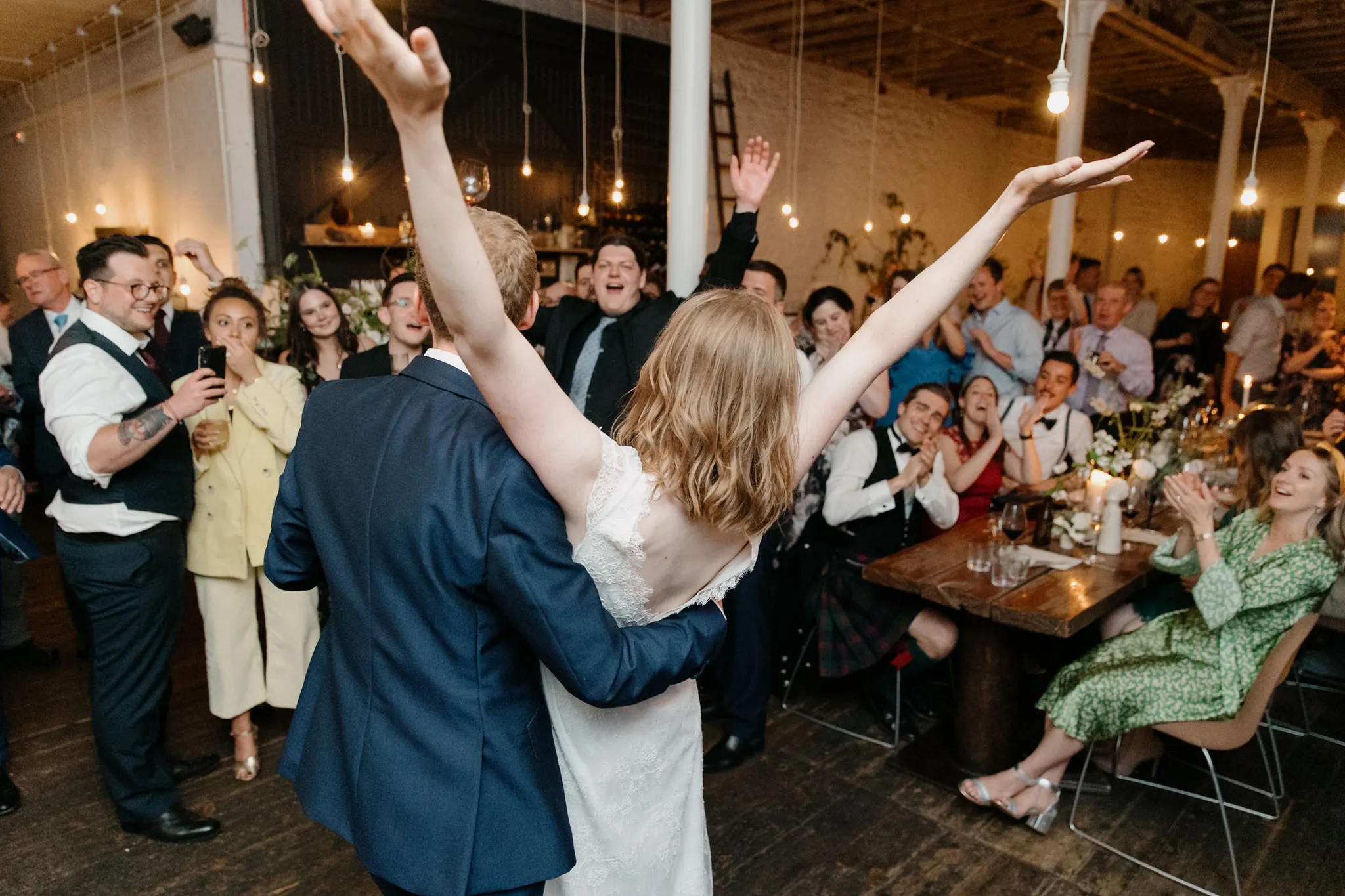 Bride and groom enter reception with arms raised as cheering guests applaud in warmly lit industrial venue