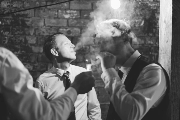 Groom in white shirt looks upward at night, bride with a cigarette — stone wall, vintage mood