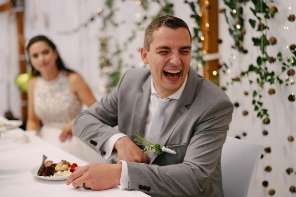 Groom laughing joyfully while eating food at wedding reception with bride nearby and green ivy decor.