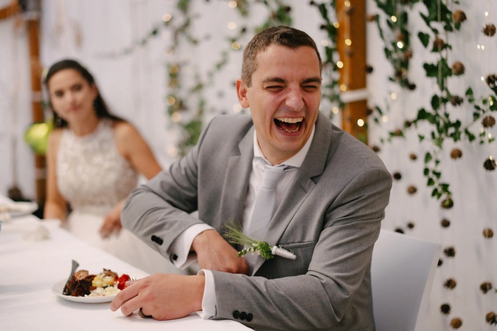 Groom laughing joyfully while eating food at wedding reception with bride nearby and green ivy decor.