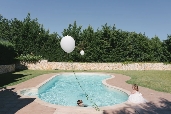 Bride in white plays at the pool edge with white balloons and a little girl in the water