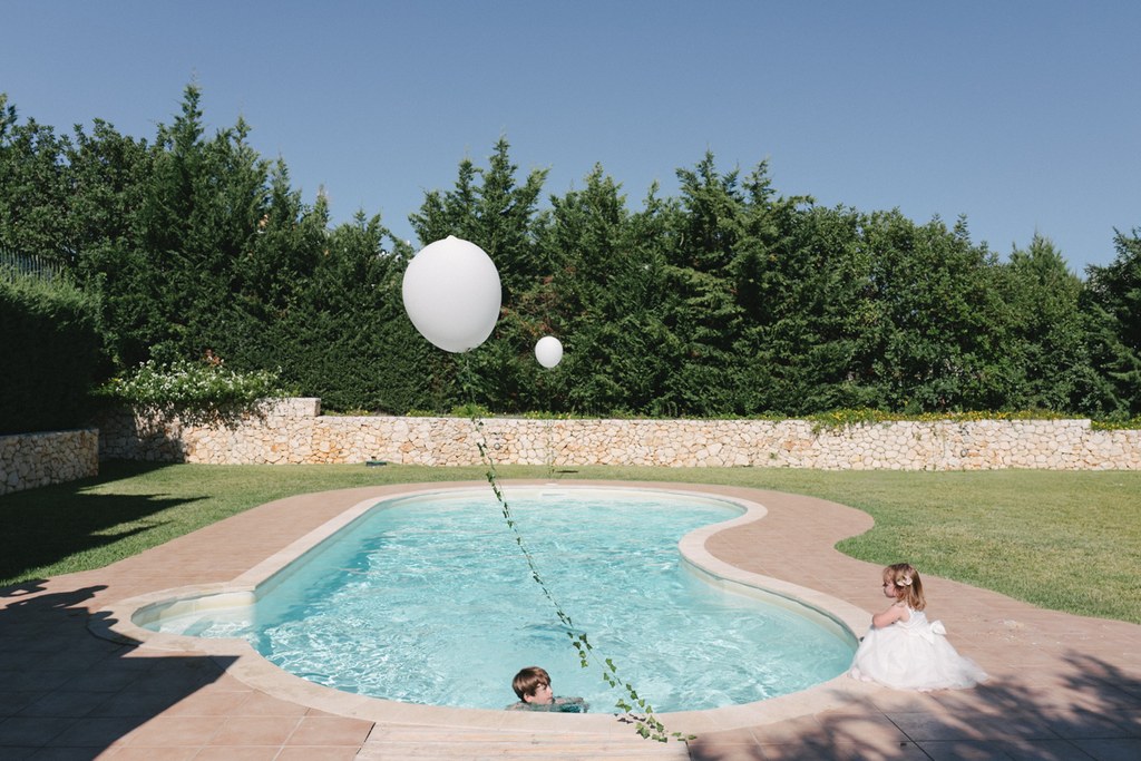 Bride in white plays at the pool edge with white balloons and a little girl in the water