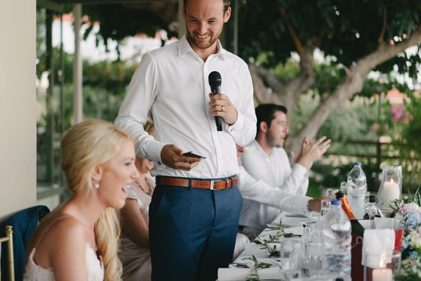 Man in white shirt and blue trousers speaks into microphone — blonde woman beside him, green garden