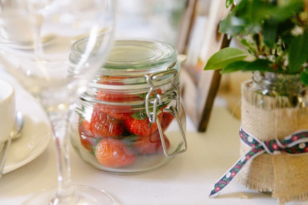 Close-up detail of a glass kilner jar filled with fresh strawberries on the wedding reception table beside a hessian-wrapped plant pot. Rustic table decor detail.