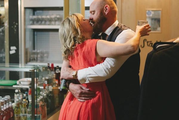 Woman in reddish dress kisses a bearded man by the bar — glass in hand, wedding celebration