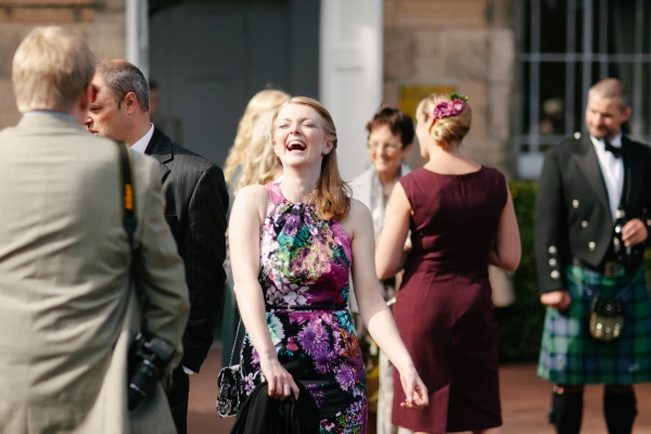 A wedding guest in a colourful floral dress laughs openly among other guests outside, including a man in a kilt. Candid documentary moment at the Royal Botanic Garden Edinburgh.