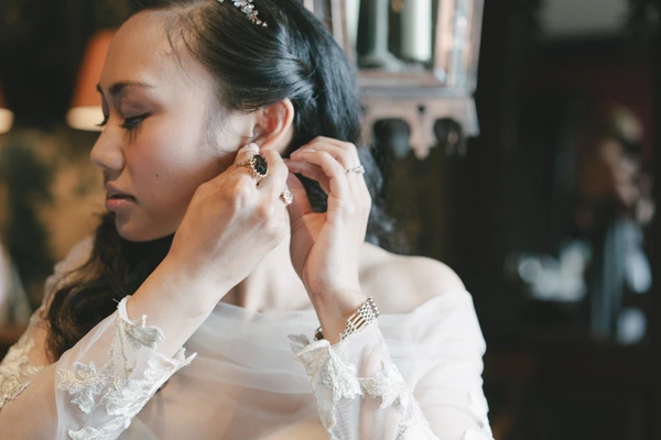 Bride in white lace carefully puts on an earring — jewels on her hand, focused expression