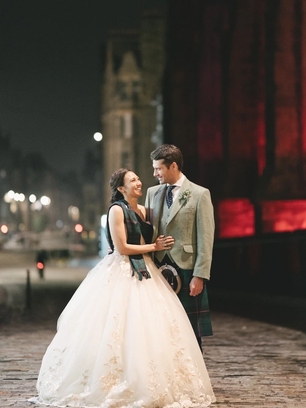 Young couple smiling at each other in a night cityscape — white lace, grey jacket and tartan kilt