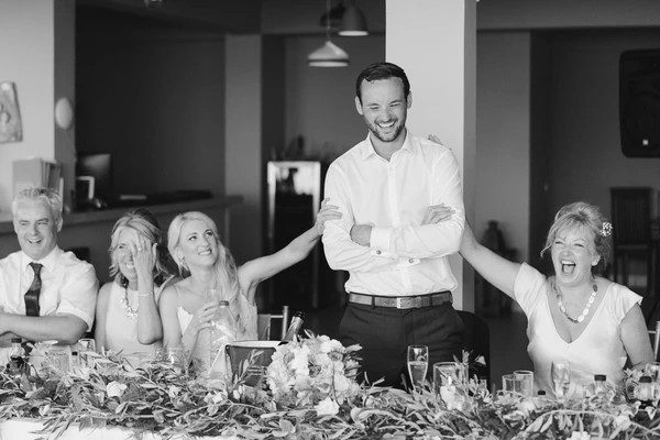Groom stands grinning with arms crossed at the table, relatives beside him laughing and listening