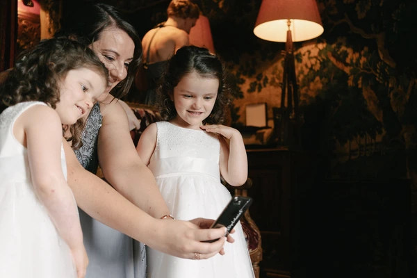 Mother and two daughters looking at a phone in warm lamp light — girl in white dress, happy smile