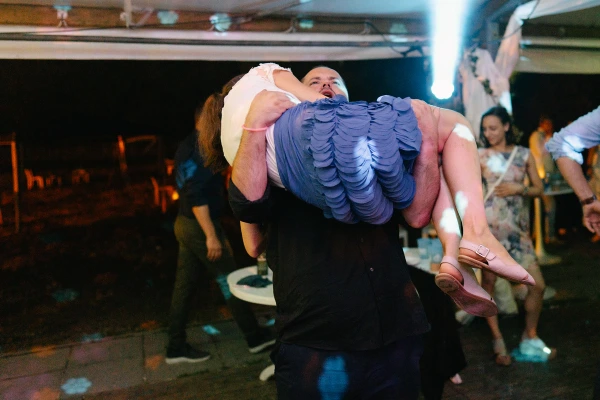 Man lifting woman joyfully at wedding celebration — colorful ambient lighting, playful dance moment, festive reception.
