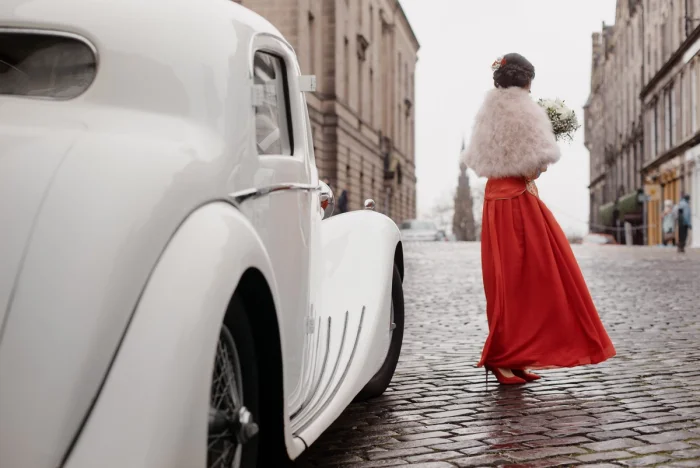 Bride in red gown and white fur wrap holds bouquet beside vintage white car on misty cobblestone street
