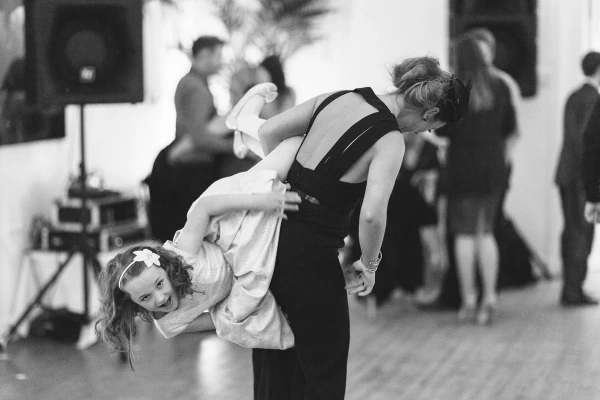 Black and white image of a woman in a black backless dress swinging a flower girl upside down on the dance floor as guests dance in the background. Energetic candid documentary wedding photograph.