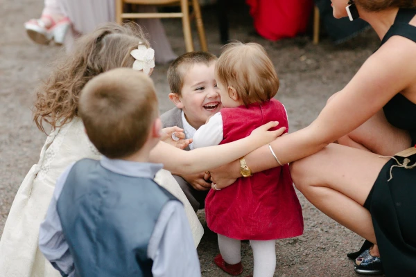 A group of young children at the wedding, one small boy beaming as he hugs a baby girl in a red dress while others gather around. Candid documentary wedding photography.