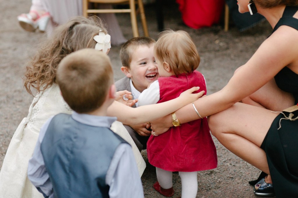 A group of young children at the wedding, one small boy beaming as he hugs a baby girl in a red dress while others gather around. Candid documentary wedding photography.