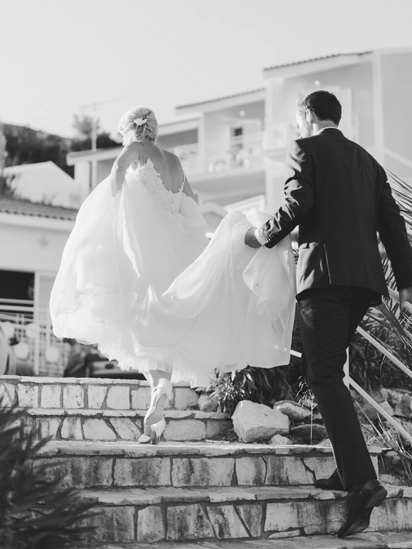 Groom in black suit helps bride on cobbled steps — white gown, elegant updo, floating lace