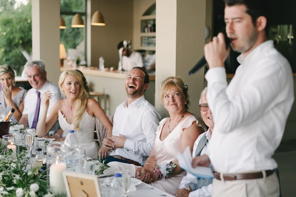 Groom gives a speech at the wedding dinner, guests laughing — bride watching happily, natural light