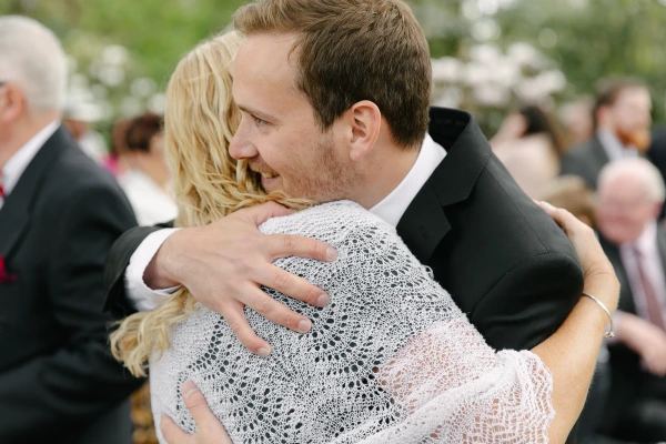 Candid shot of the groom hugging a smiling blonde guest after the ceremony, her eyes closed in the embrace. Warm post-ceremony candid documentary photograph.