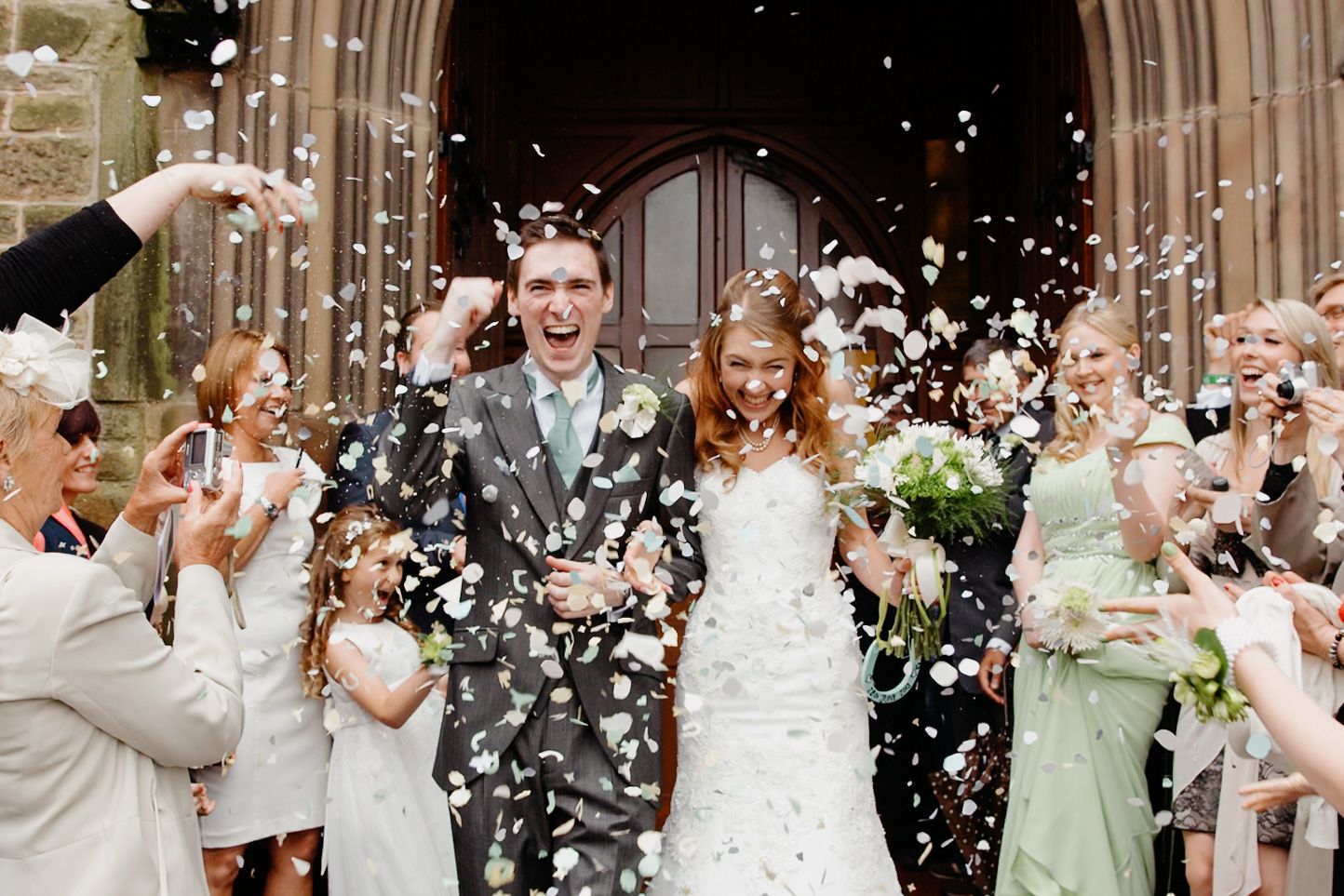 Joyful newlyweds exit stone church amid shower of white confetti, surrounded by cheering guests in a sunny outdoor celebration.