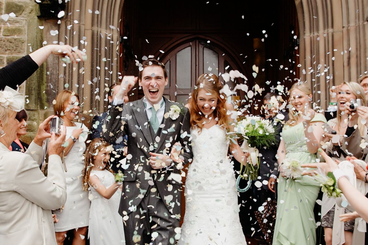 Joyful newlyweds exit stone church amid shower of white confetti, surrounded by cheering guests in a sunny outdoor celebration.