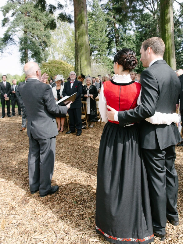 Bride and groom exchanging rings during the outdoor ceremony while the officiant guides them, guests and gardens visible in the background. Candid ring exchange moment.