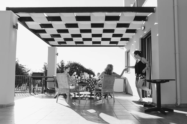 Woman serves a couple on a terrace — checkerboard pergola, long shadows in warm sunlight. Black-and-white.
