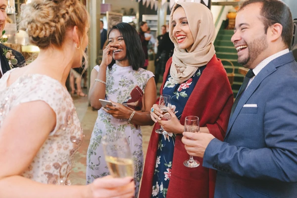 Bride in lace smiling among guests — woman in hijab, suited man, guests with glasses in hand