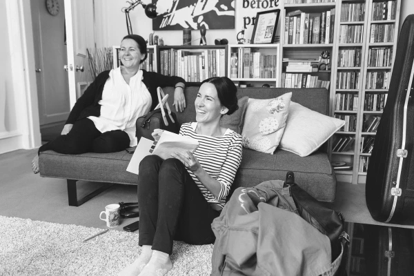 Black and white candid shot of two women relaxing at home, one reading on the floor and one seated on a sofa, surrounded by bookshelves. Relaxed bridal morning preparations.