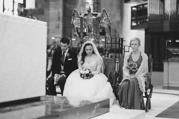 Bride and groom in the church pew with witness, church sculpture visible behind — black-and-white