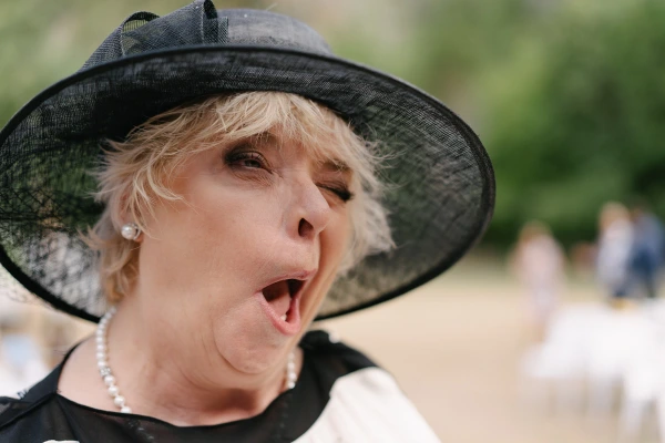 Senior guest in navy hat expressing joyful emotion with animated expression at wedding outdoor event.
