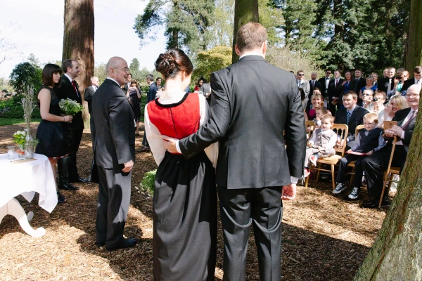 Back view of the bride and groom standing together at the outdoor altar, guests seated among giant redwood trees of the Royal Botanic Garden Edinburgh. Wide ceremony shot.