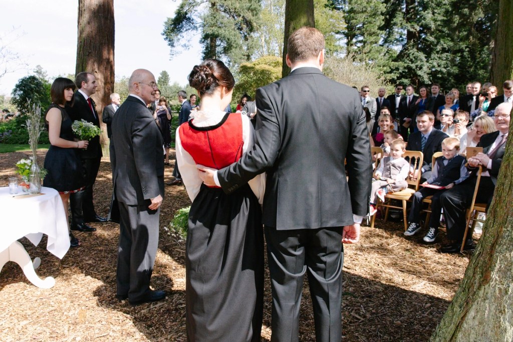 Back view of the bride and groom standing together at the outdoor altar, guests seated among giant redwood trees of the Royal Botanic Garden Edinburgh. Wide ceremony shot.