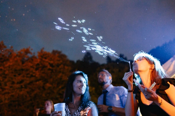 Wedding guests blowing bubbles outdoors at night — evening celebration, magical bokeh lights, festive party atmosphere.