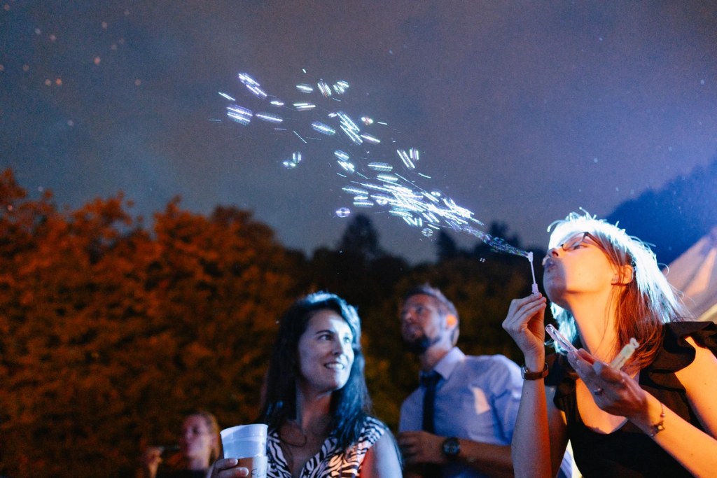 Wedding guests blowing bubbles outdoors at night — evening celebration, magical bokeh lights, festive party atmosphere.