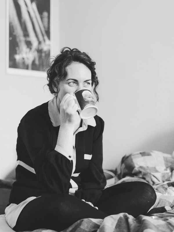 Woman in black suit on the bed holding a coffee cup, pensive gaze — black-and-white portrait