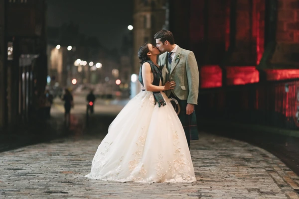 Newlyweds kiss at midnight — ivory tulle gown, grey suit with tartan, red neon and city lights behind