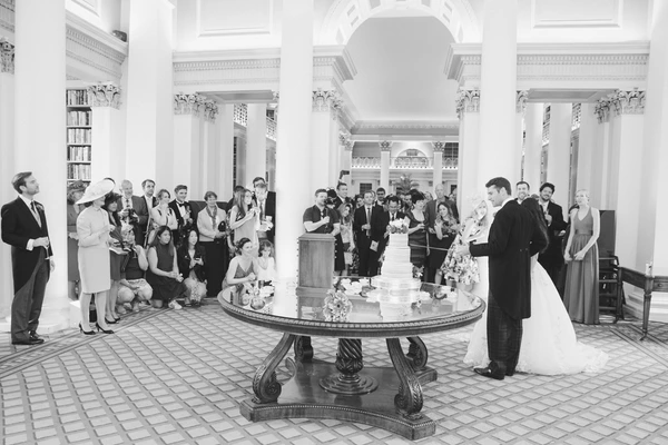 Bride stands beside the decorated cake in a white hall, guests toast with glasses