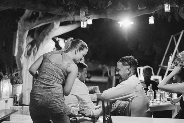 Woman dances between two men at the night party — fairy lights in branches, tables and guests around