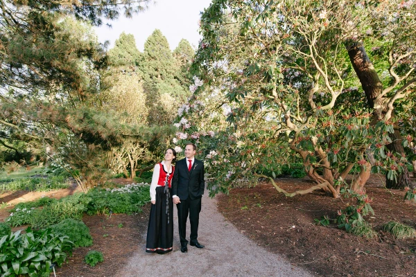 The bride in a Norwegian bunad and the groom in a dark suit stand together on a garden path among towering trees and flowering rhododendrons at the Royal Botanic Garden Edinburgh. Documentary wedding portrait.